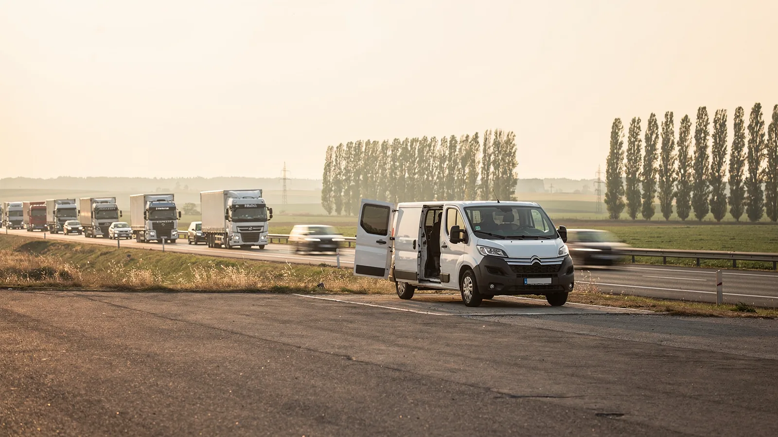 Bijeli kombi s otvorenim bočnim vratima parkiran na proširenju uz kolonu kamiona, ravnica i topole u pozadini